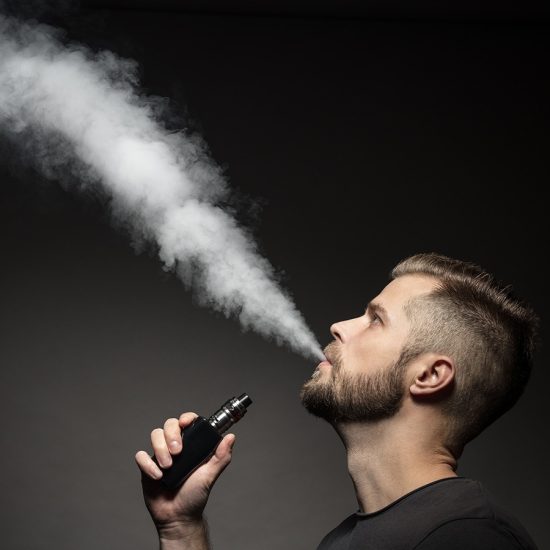 A side profile of a man blowing a smoke of a vape upward.
